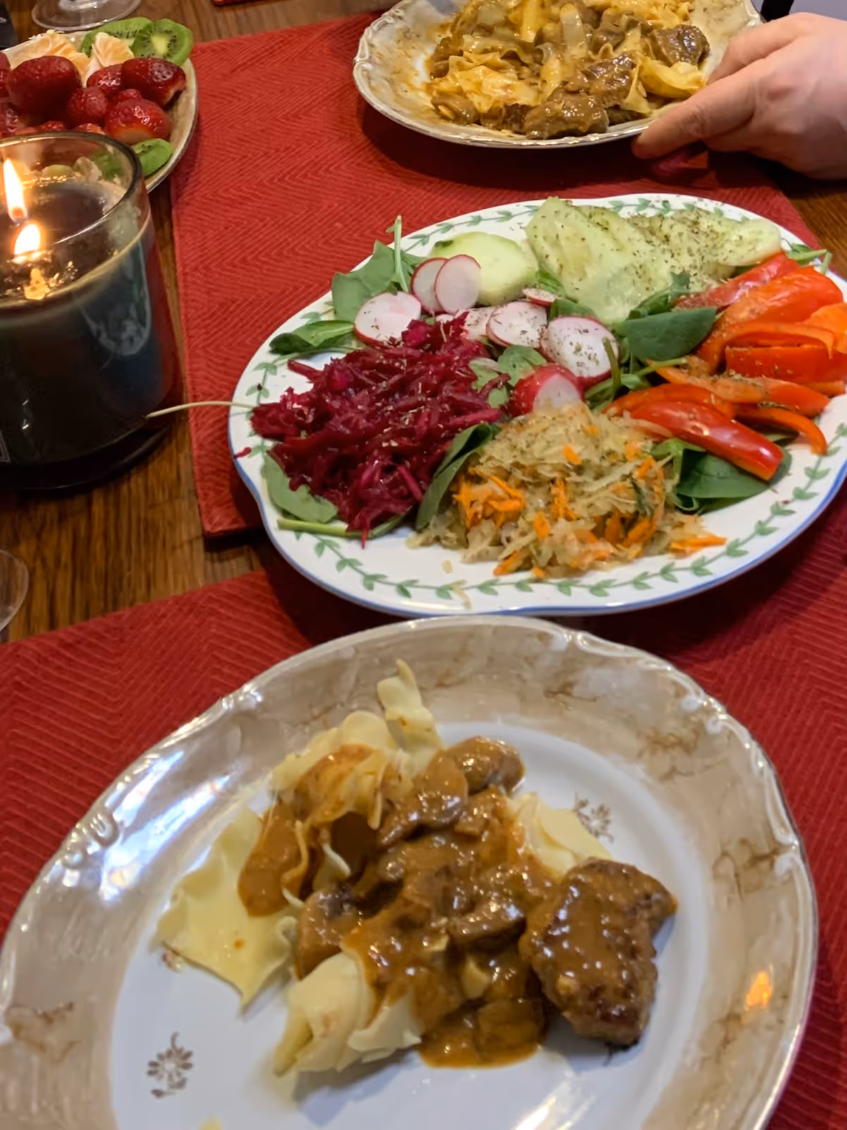 Table setting with a large salad plate, meat dish, and candle on a red tablecloth.