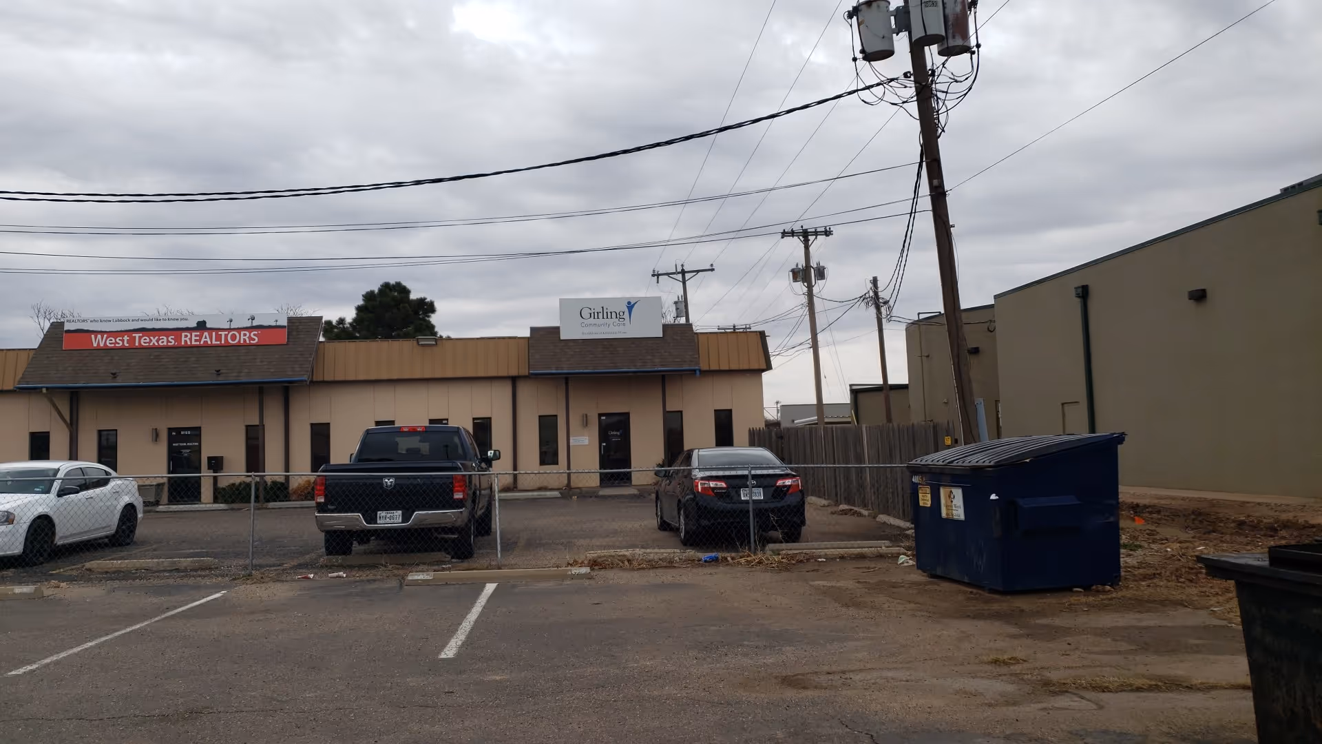 Exterior view of a single-story facility with a parking lot, fence, and dumpsters.