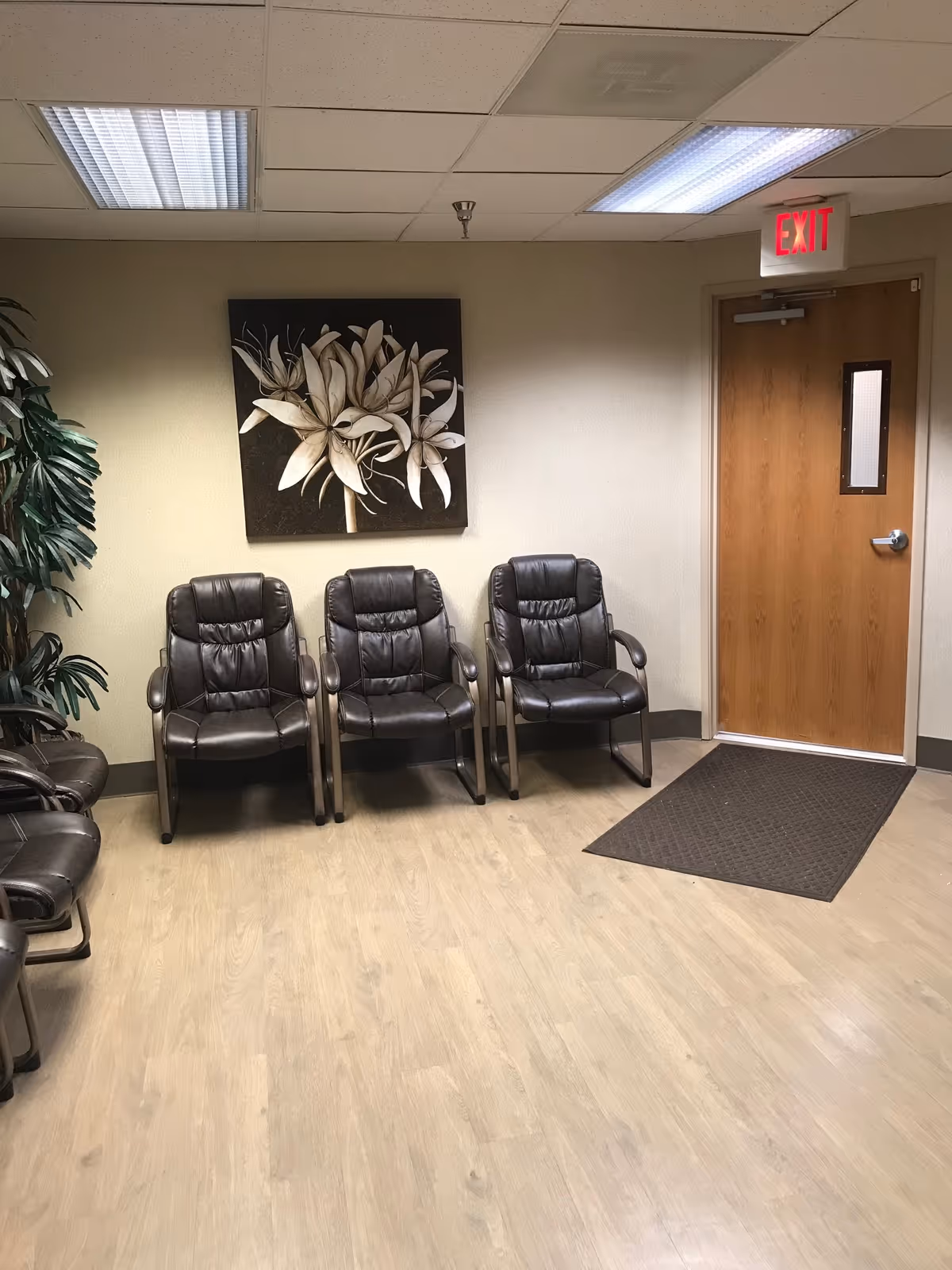 Lobby waiting area with black leather chairs, plant, floral artwork, exit door, and wooden flooring.
