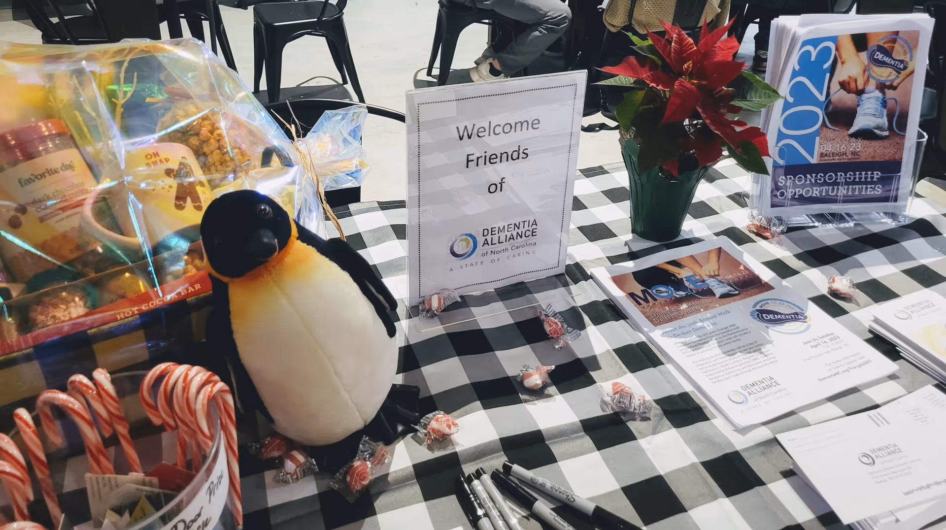 Promotional table setup with a penguin plush, candy, brochures, and a welcome sign indoors.