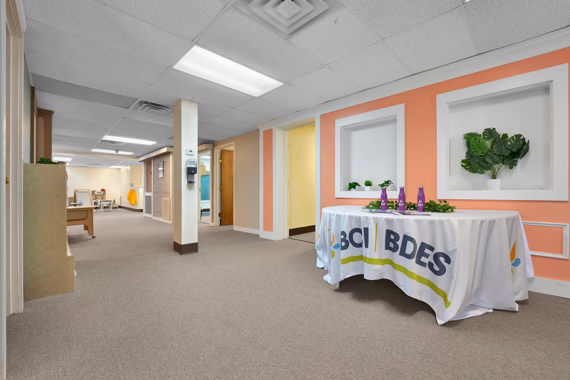 Indoor hallway with a branded table and potted plants along an orange-walled corridor