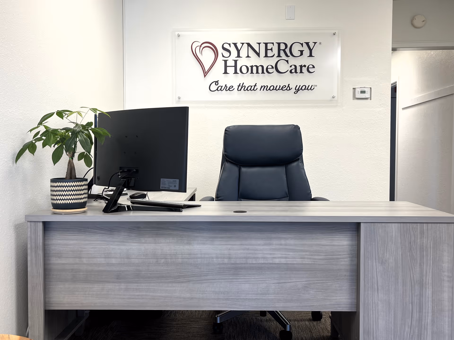 Reception desk with computer, potted plant, and a Synergy HomeCare sign on the wall.