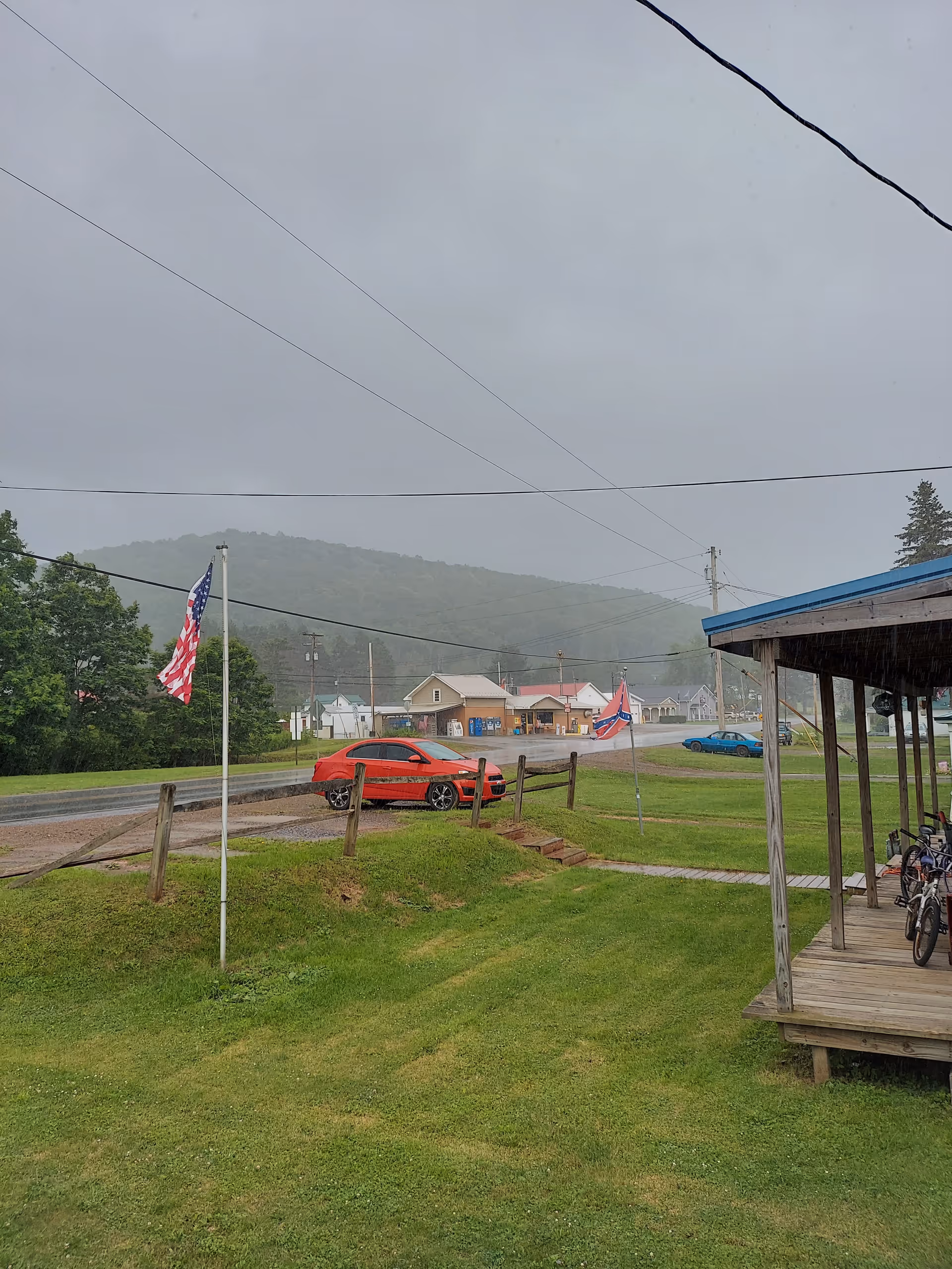A rural outdoor scene with grassy yard, American flag, cars, and a covered porch.