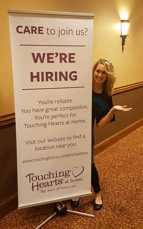 Smiling woman stands beside a WE'RE HIRING banner in a hallway.