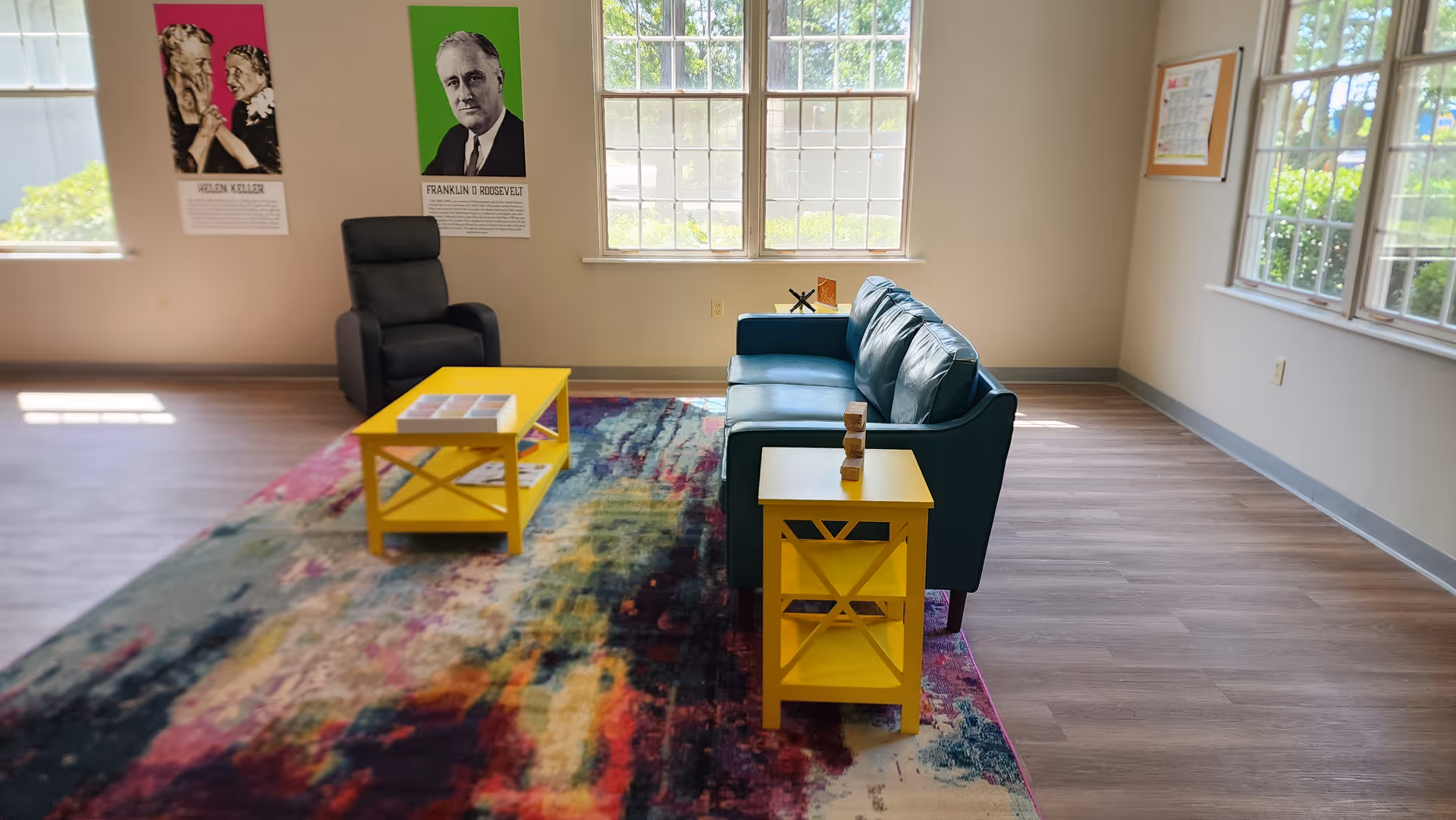 Bright senior living lounge with a colorful rug, yellow tables, teal sofa, and wall posters.