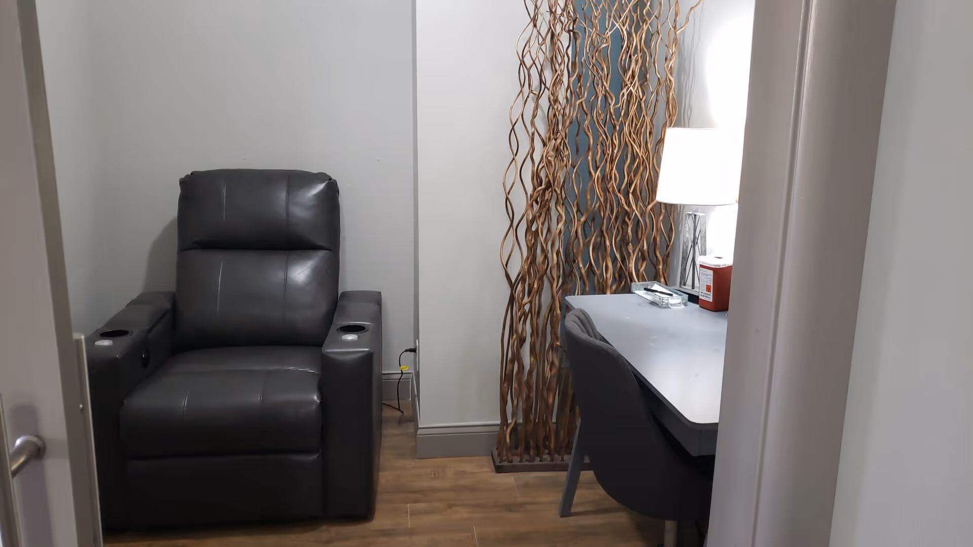 A dark gray leather recliner sits beside a desk and lamp in a small room.