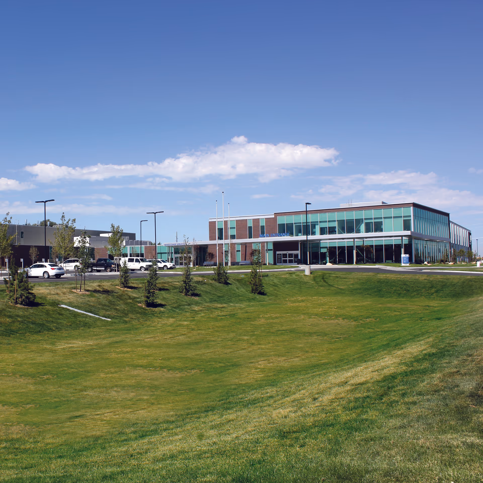 Modern brick-and-glass building with a green lawn and parked cars in front.