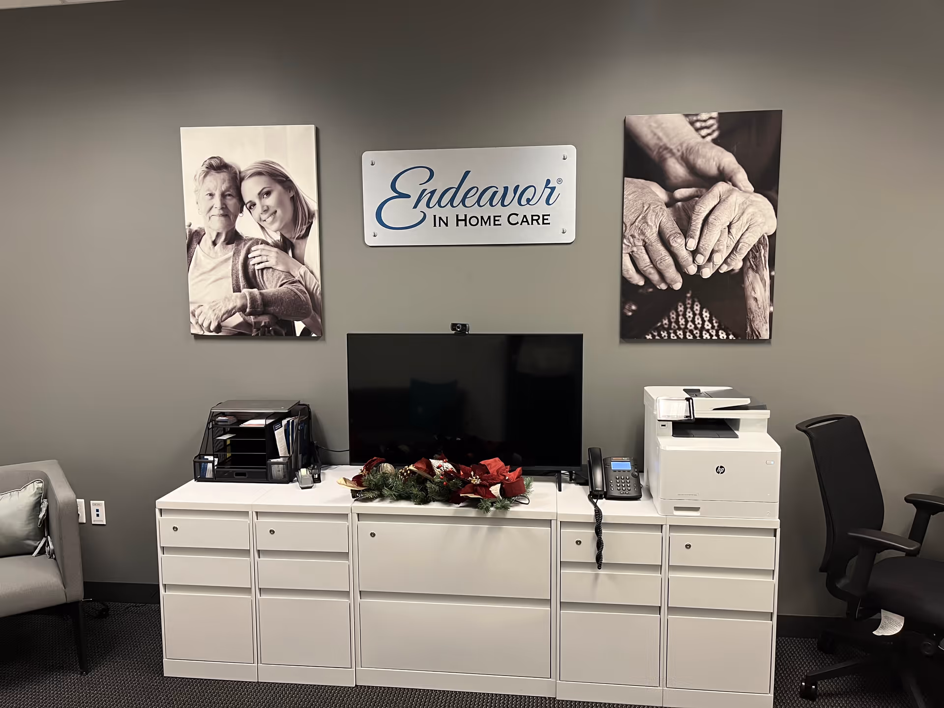 Office reception area featuring Endeavor In Home Care sign, monitor, and printer against gray wall.