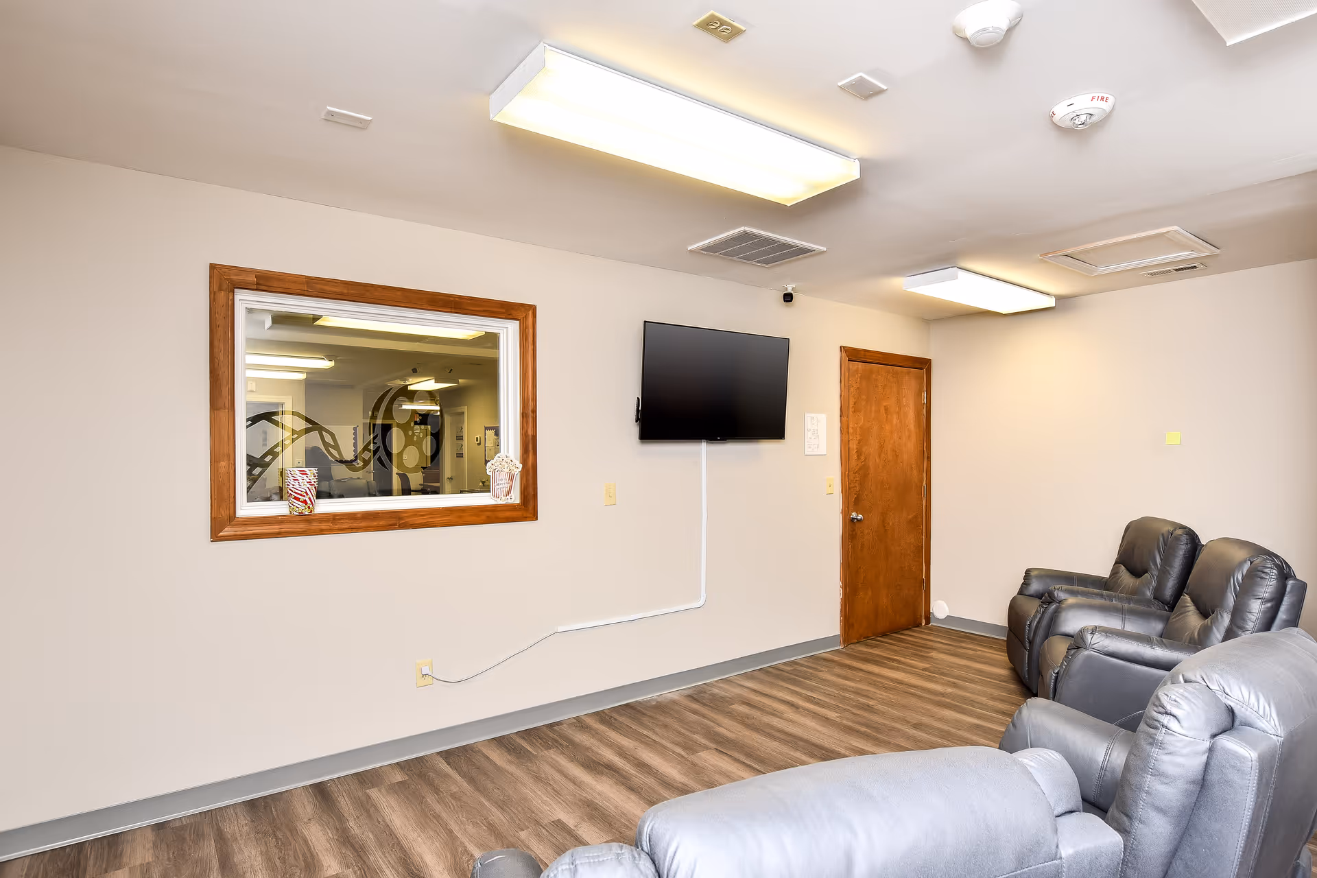 Interior senior living lounge with beige walls, wall TV, door, and recliner chairs.