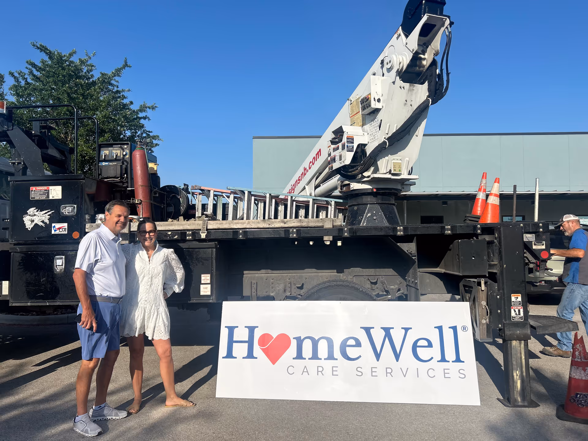 Outdoor scene with a large crane truck, two people, and a HomeWell Care Services banner.