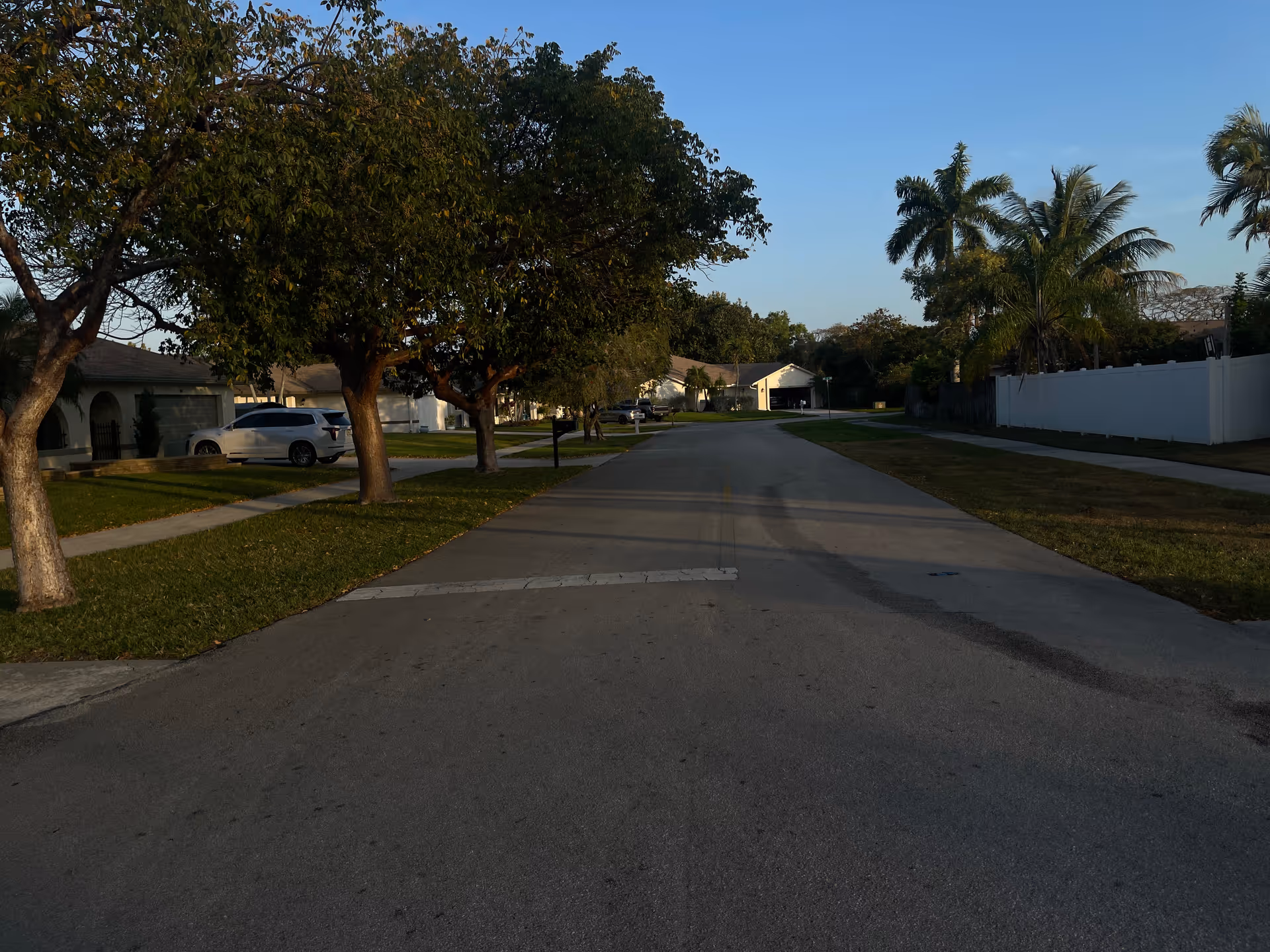 Quiet residential street with trees, sidewalks, and a white fence along single-family homes.