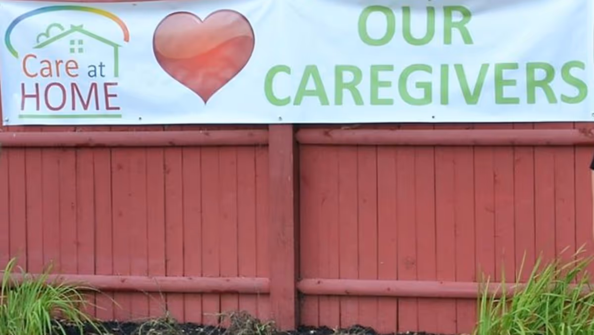 Care At Home LLC banner with a heart reads Our Caregivers on a red wooden fence.