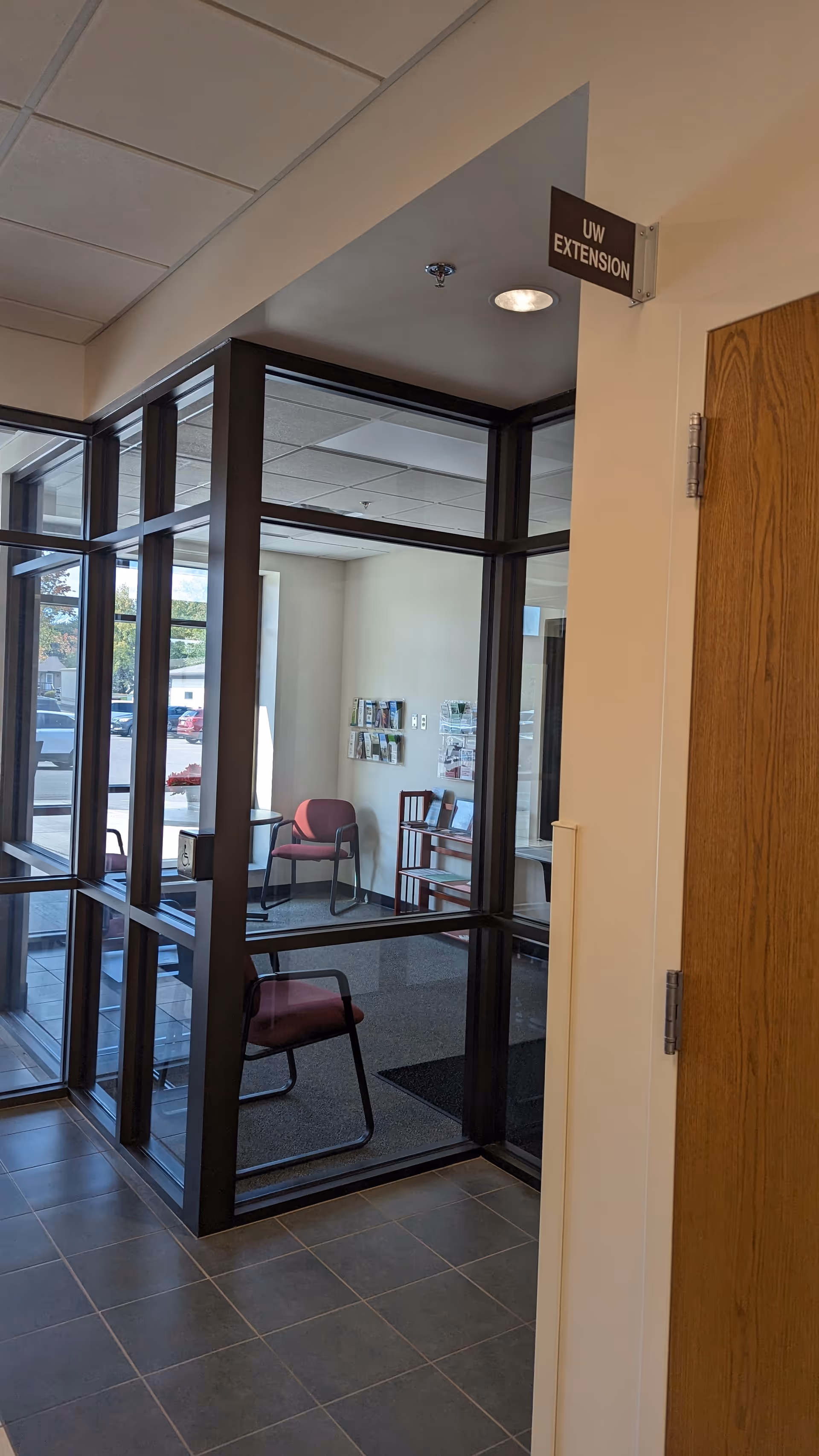 Glass-walled waiting area inside a facility with pink chairs and a pamphlet rack.