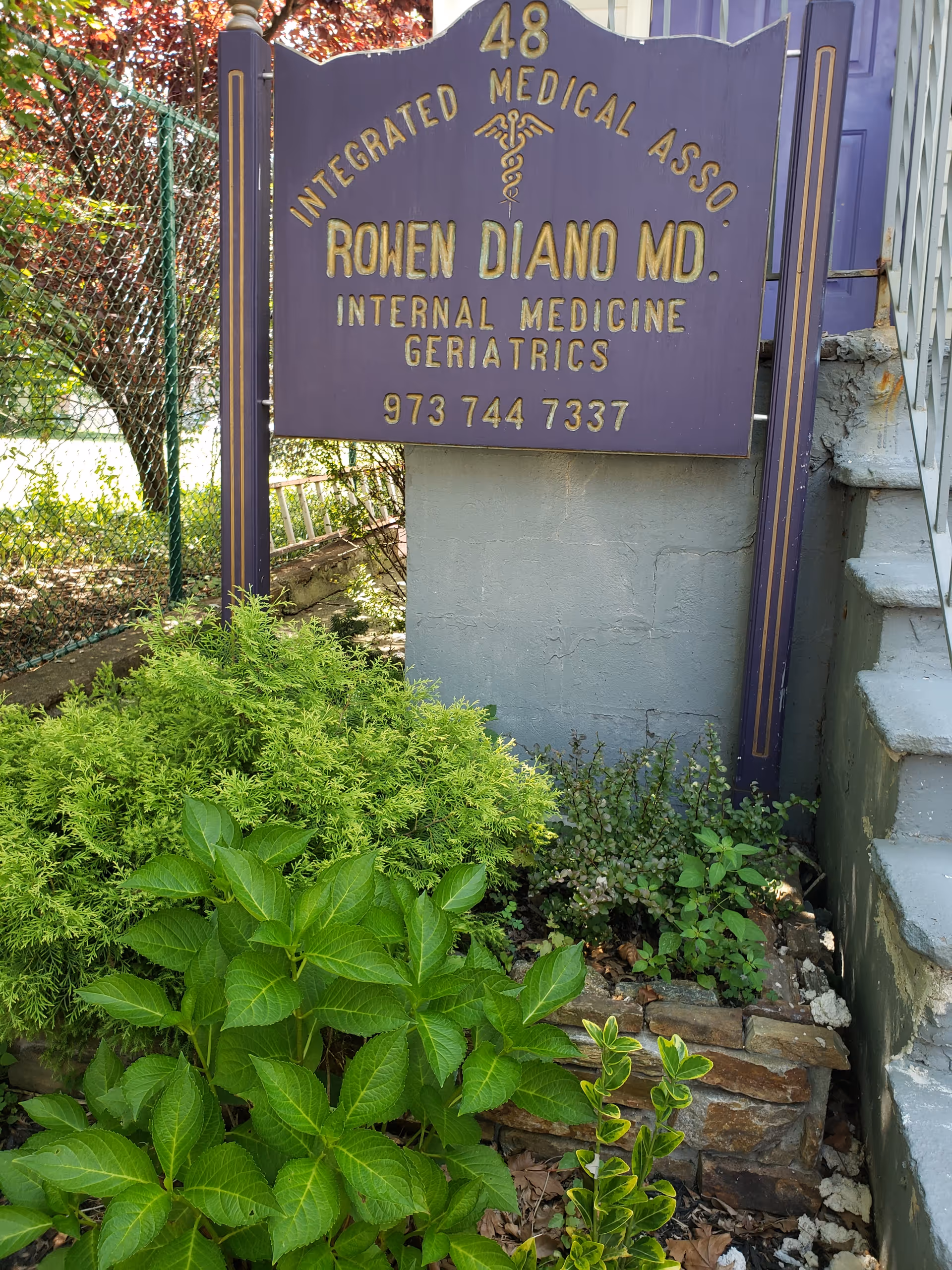 Purple medical sign on stone base beside steps with greenery outside a building
