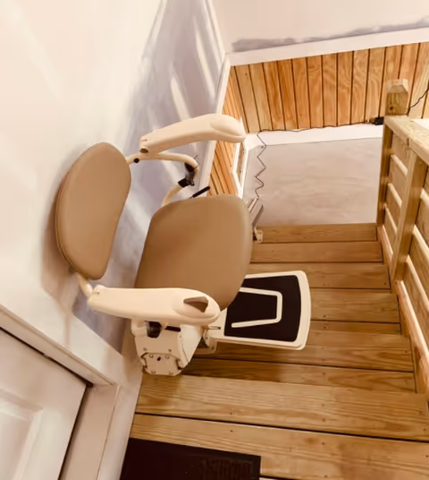 Beige stairlift chair mounted beside a wooden staircase inside a senior living facility.