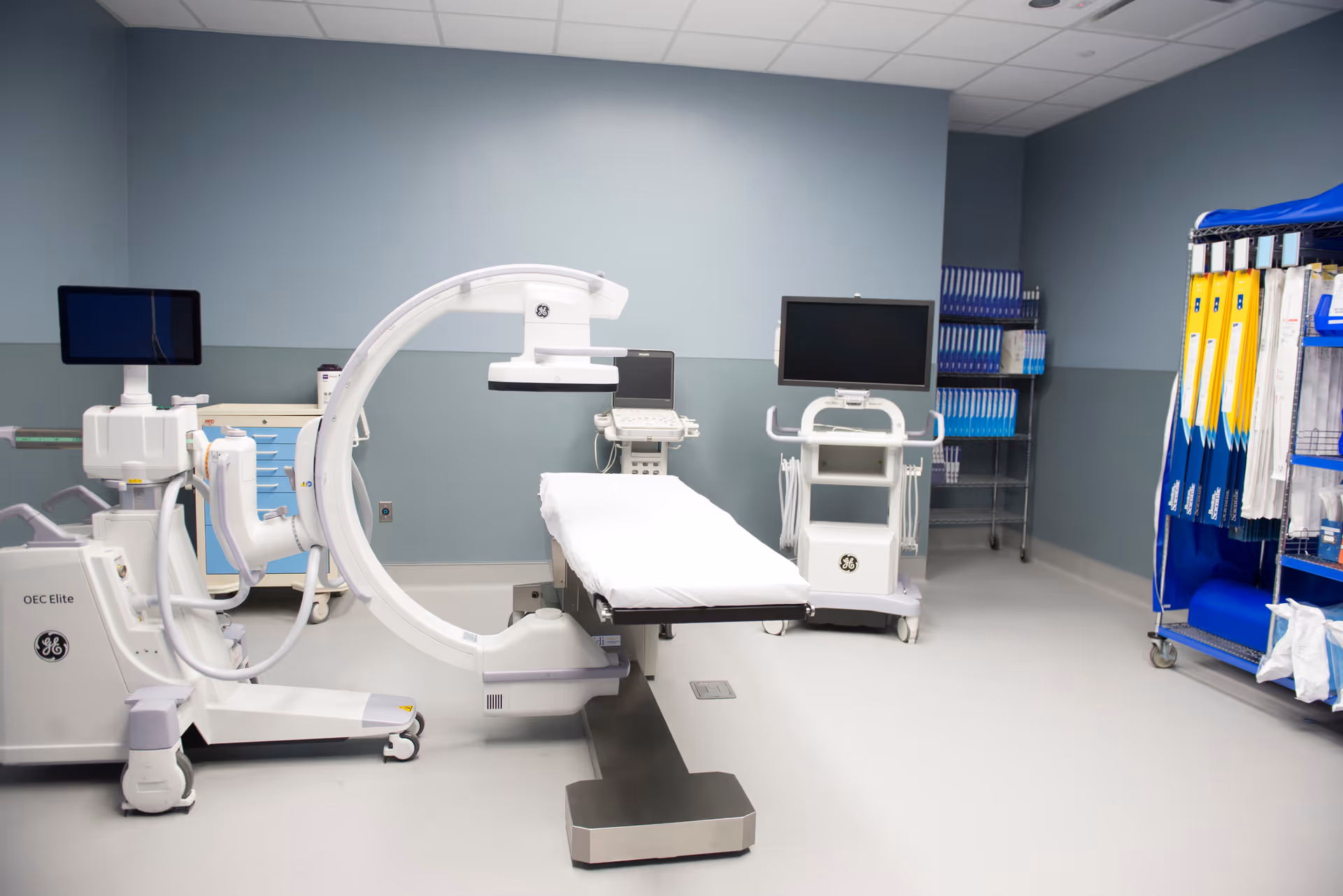 A clinical imaging room with a large medical scanner, monitors, and organized equipment against blue walls.