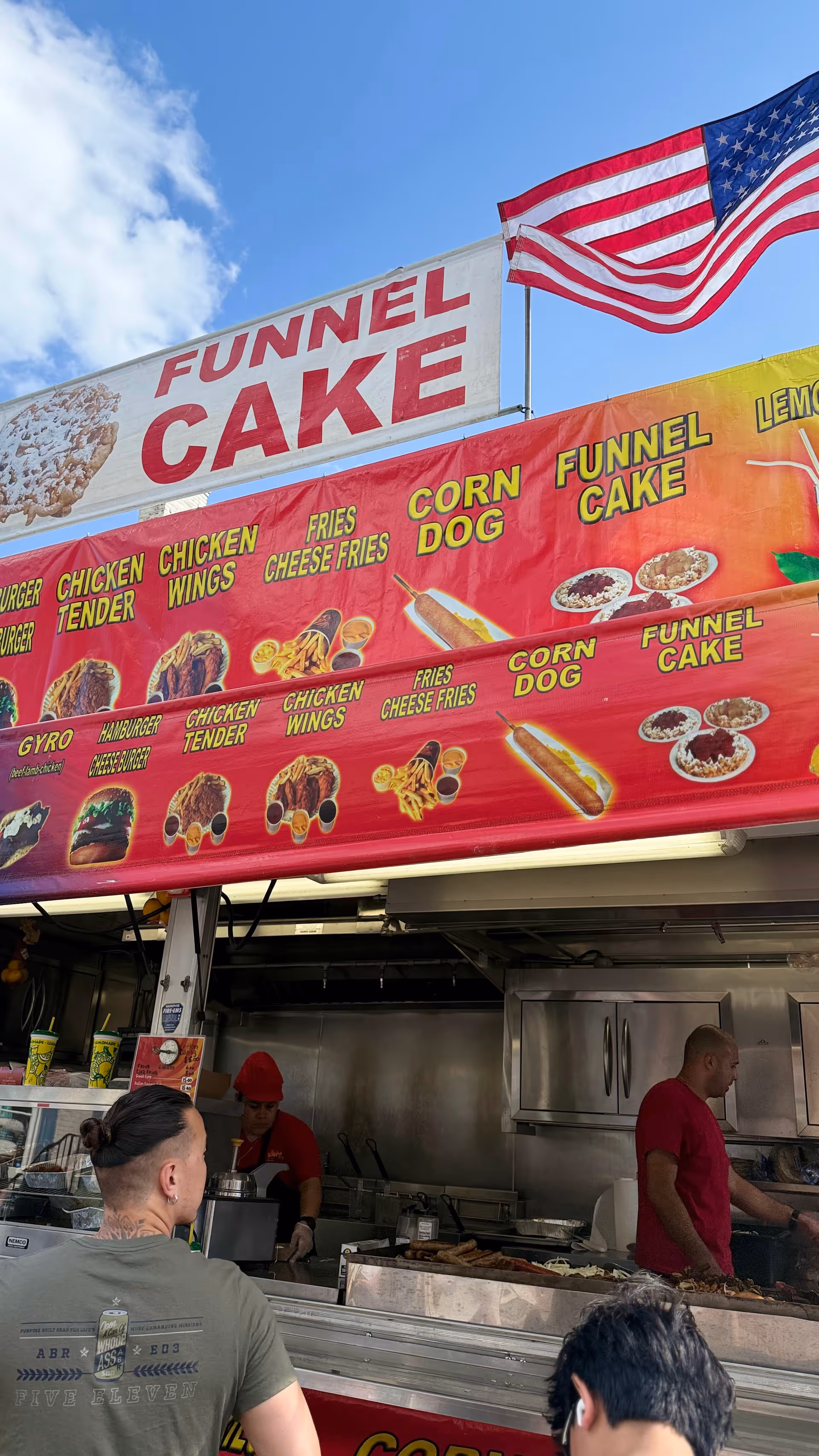 Outdoor food stall with funnel cake sign and workers cooking under an American flag