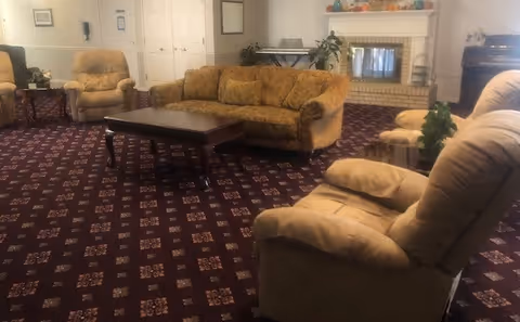 Senior living lounge with beige sofas, chairs, a coffee table, and a fireplace.
