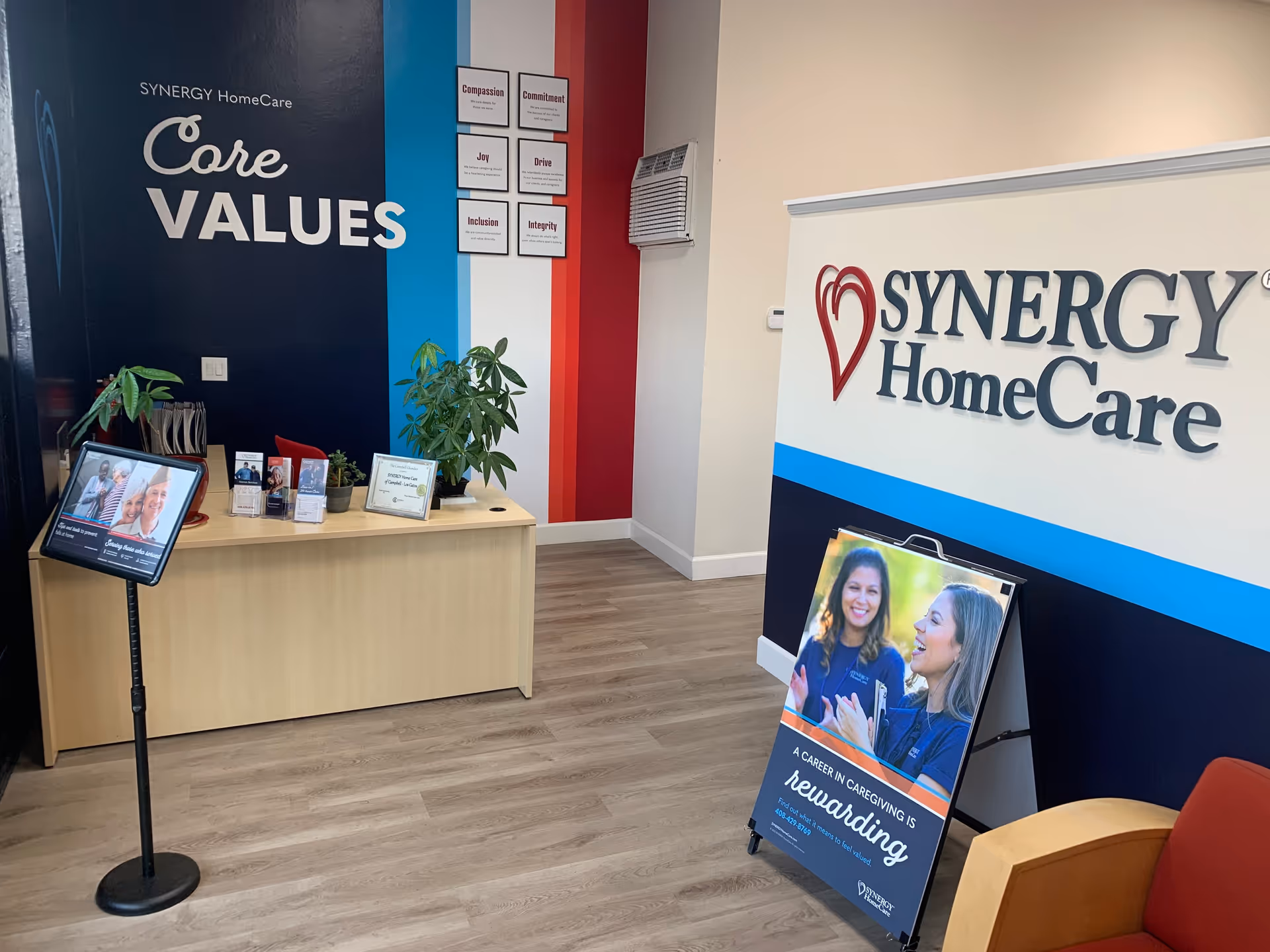 Reception area with a wooden desk, plants, and a large SYNERGY HomeCare banner.