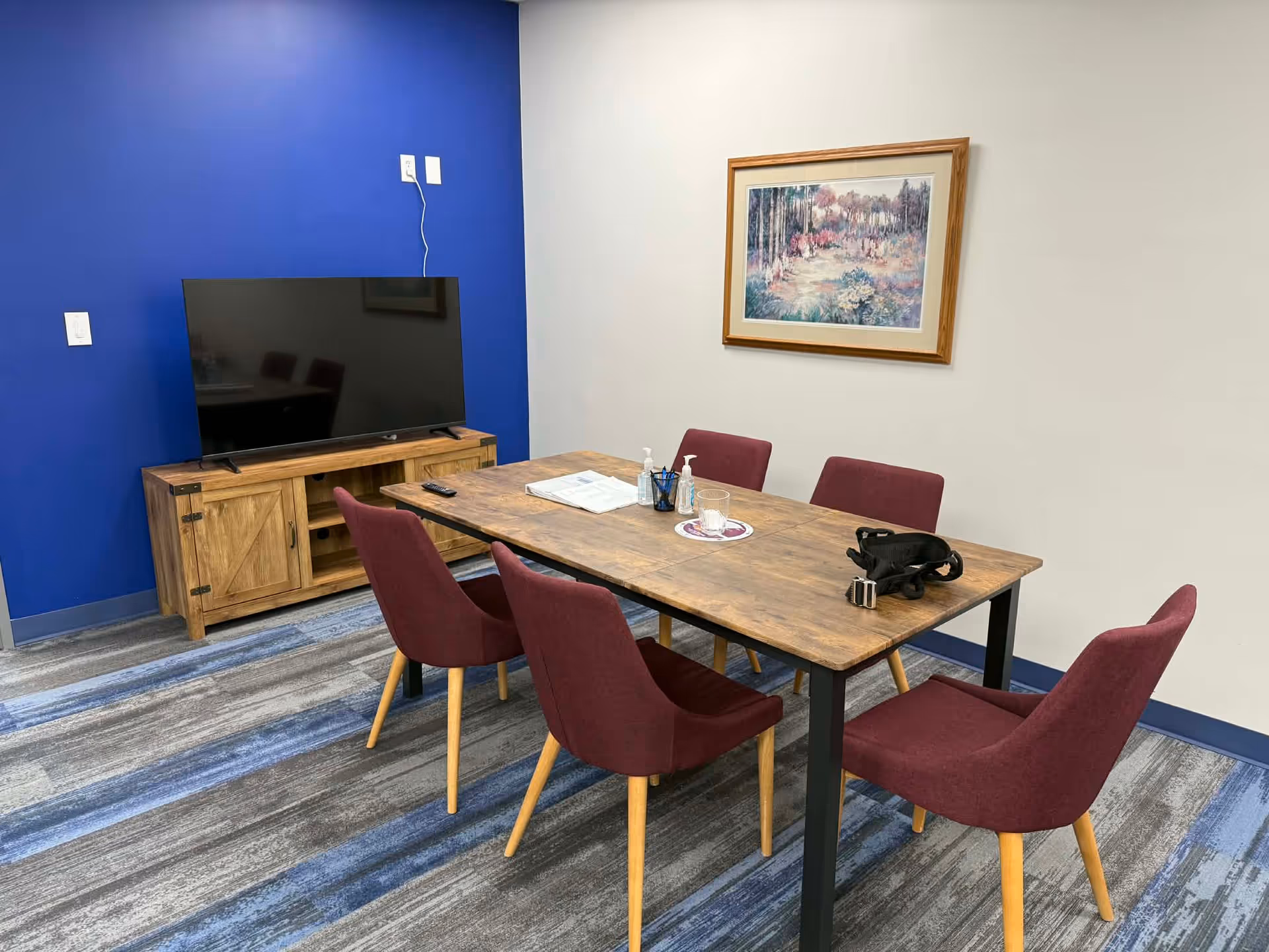 Communal dining room with wooden table, burgundy chairs, blue accent wall, and TV cabinet.