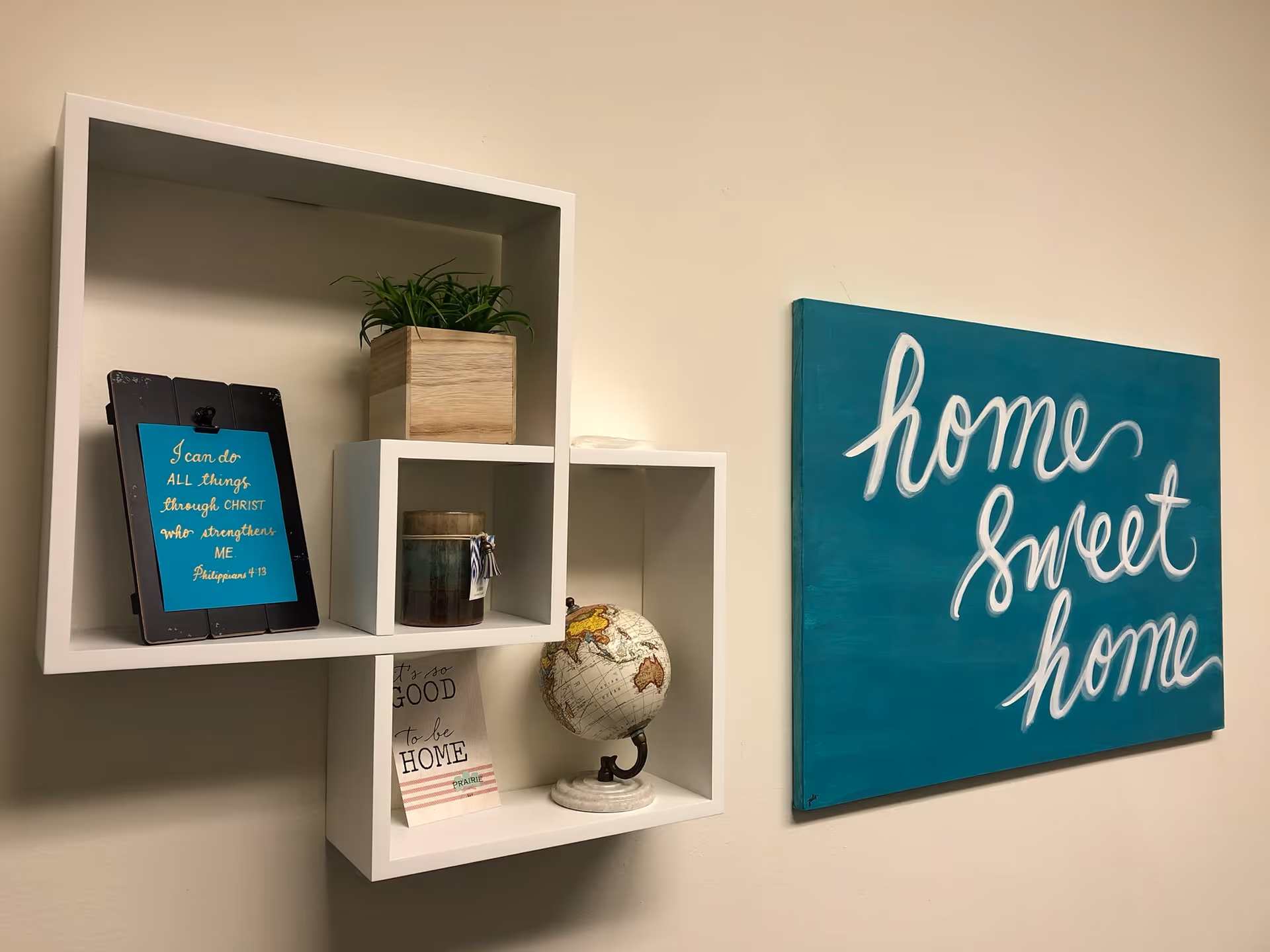 Decorative white wall shelves with plants, a globe, and teal 'Home Sweet Home' artwork.