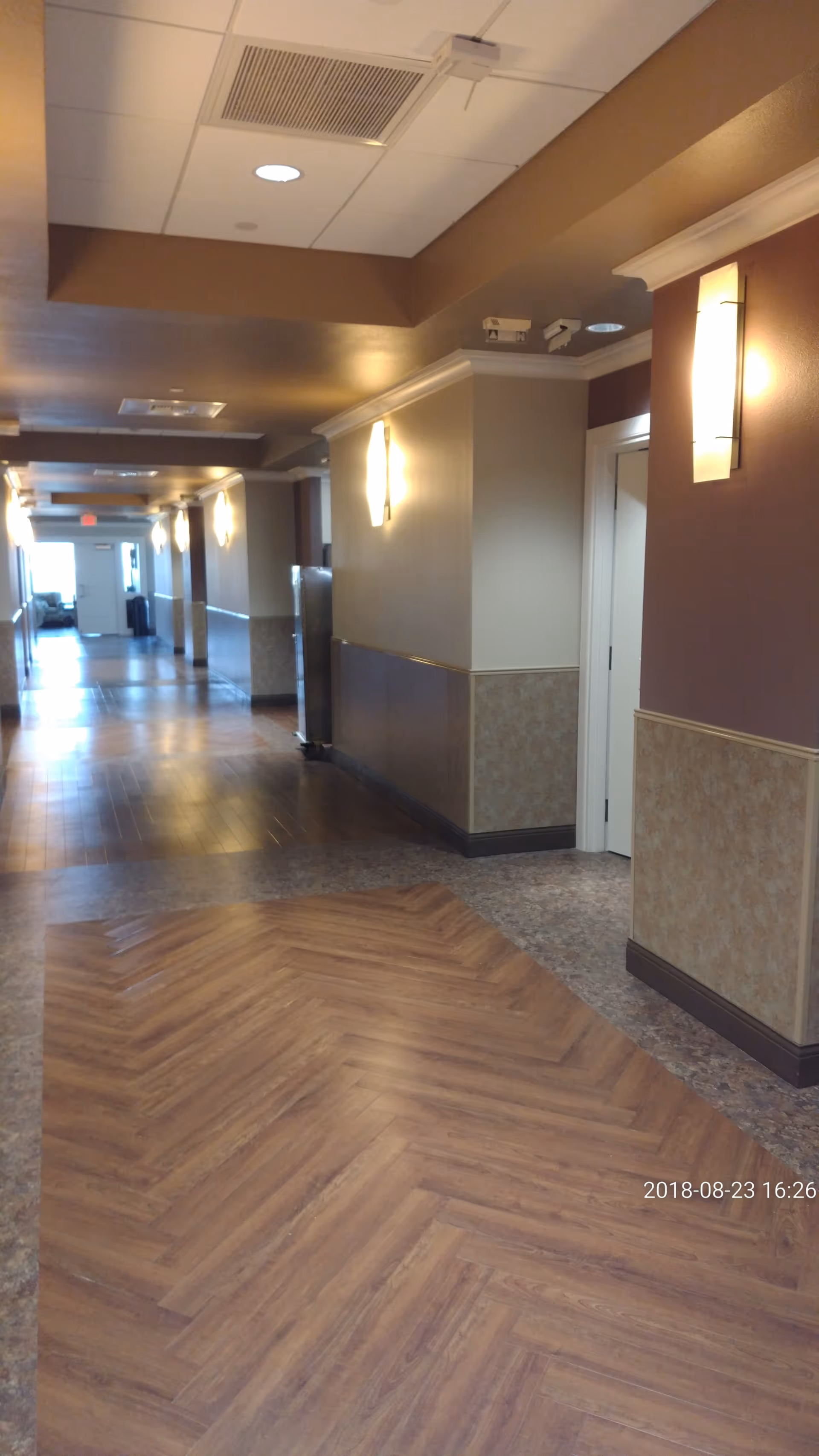 Long hallway in a senior living facility with wood-patterned flooring and wall sconces.
