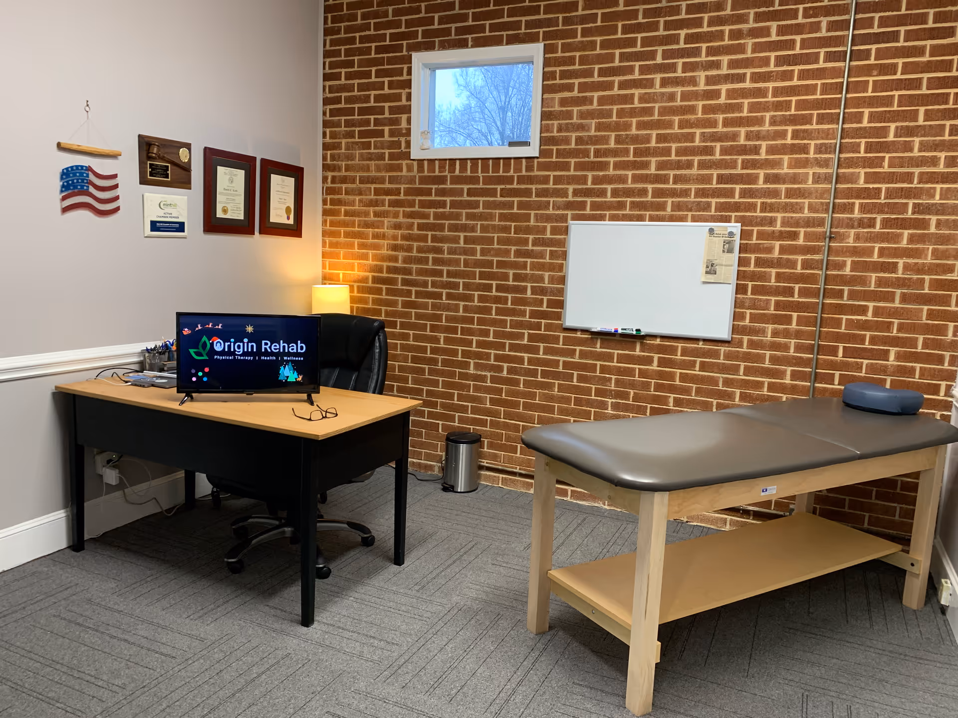 A rehab therapy office with a desk, computer, certificates, whiteboard, and treatment table.