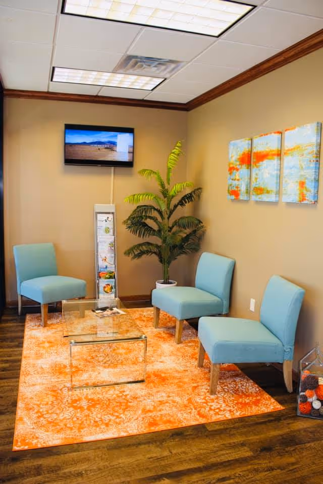 Bright waiting area with blue chairs, a plant, wall art, and a glass coffee table.