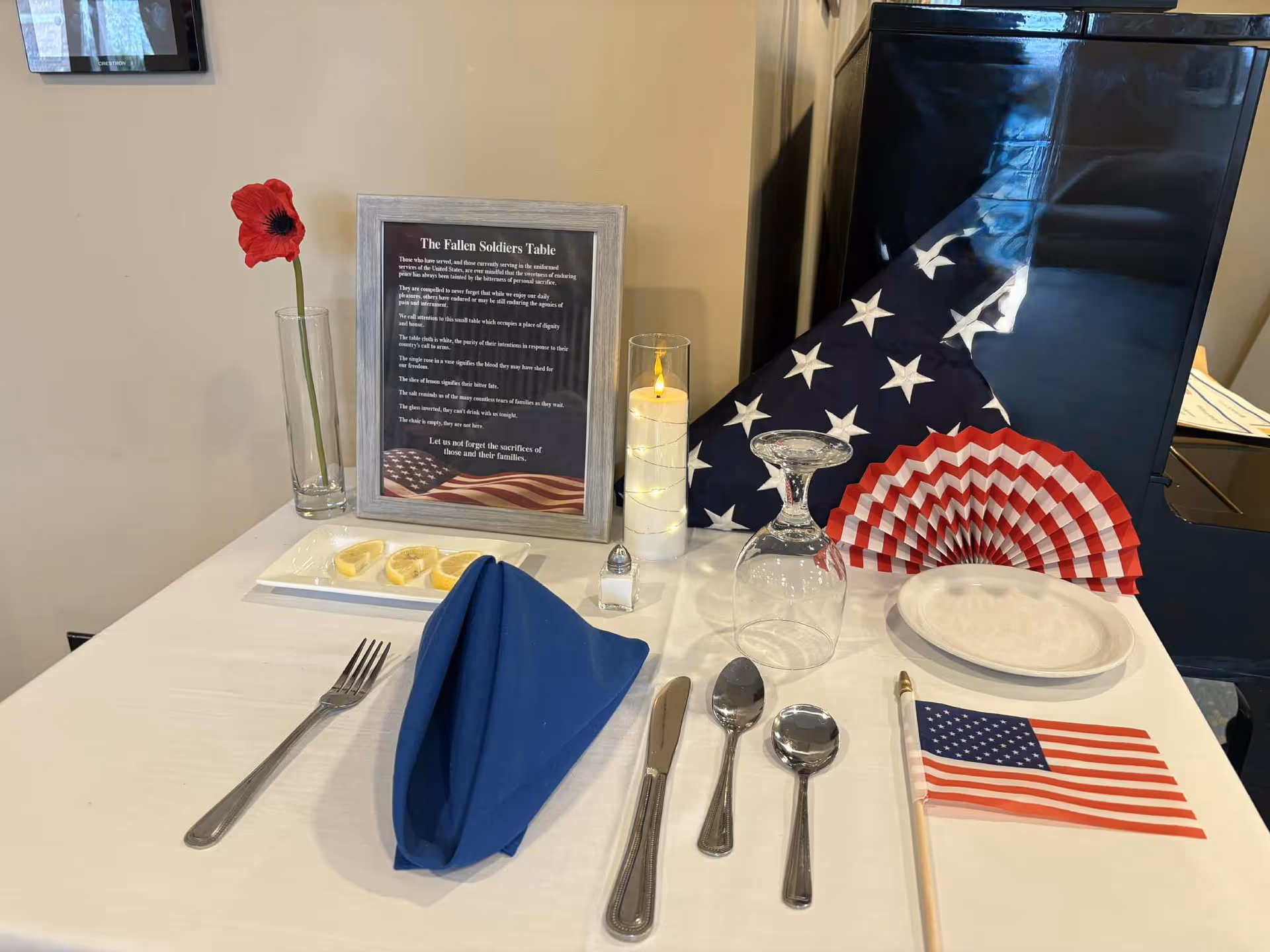 Memorial table display with a framed tribute, candle, flower, flag, and utensils on a white table.
