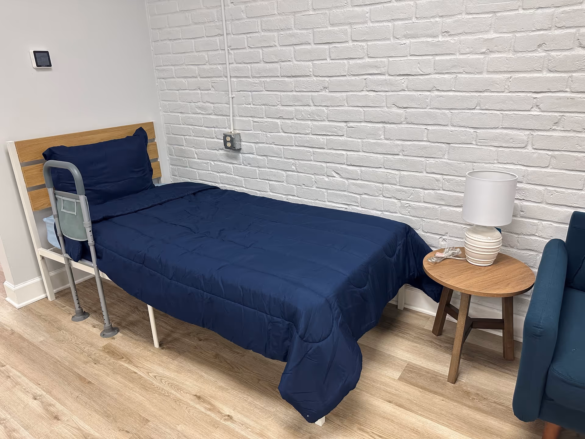Single blue bed with rail, beside round table and lamp in a small room.