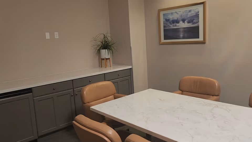 A dining room with a marble table, tan chairs, cabinets, plant, and framed wall art.