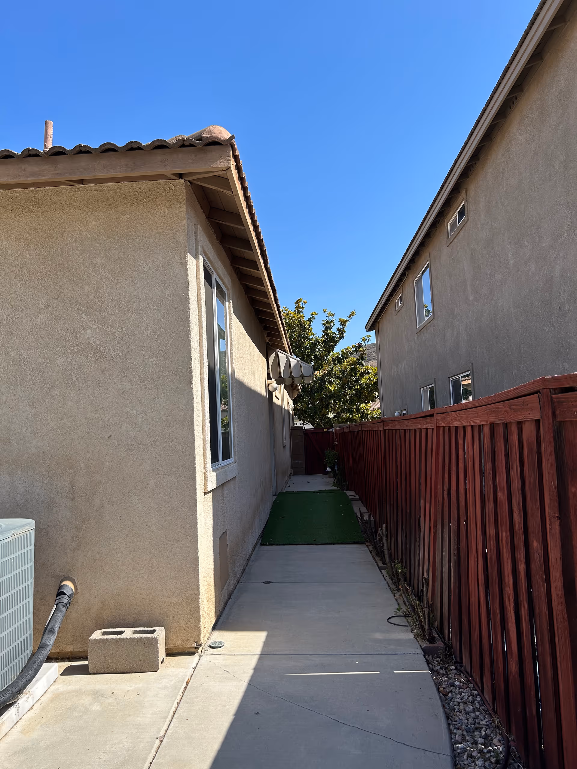 Narrow concrete walkway between two beige residential buildings with a wooden fence and an air conditioner.