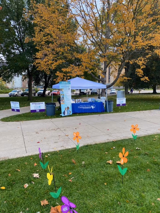 Outdoor Visiting Angels information tent with blue canopy and banners on a grassy, tree-lined campus.