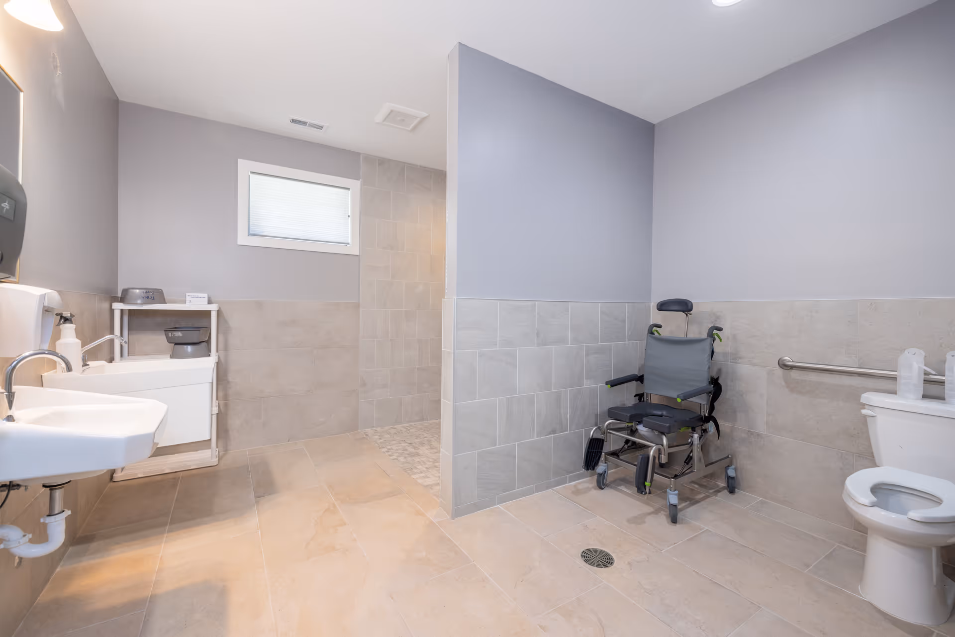 Accessible bathroom with sink, toilet, wheelchair, and grab bars in a gray-tiled space.
