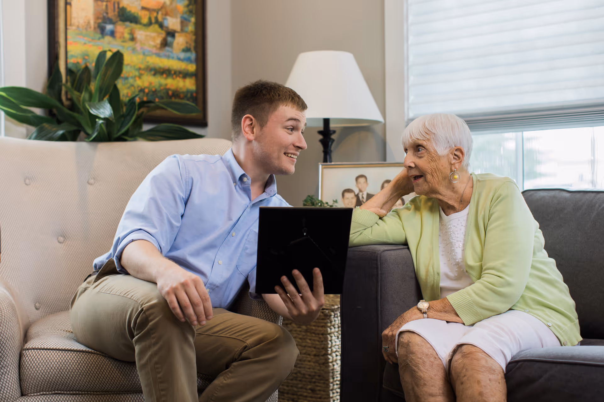 A caregiver and elderly resident converse on a living room couch in a senior facility.