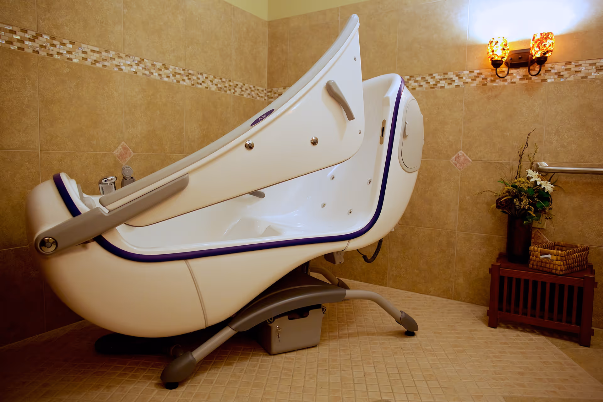 A large white treatment chair sits in a beige tiled room with a table and flowers.