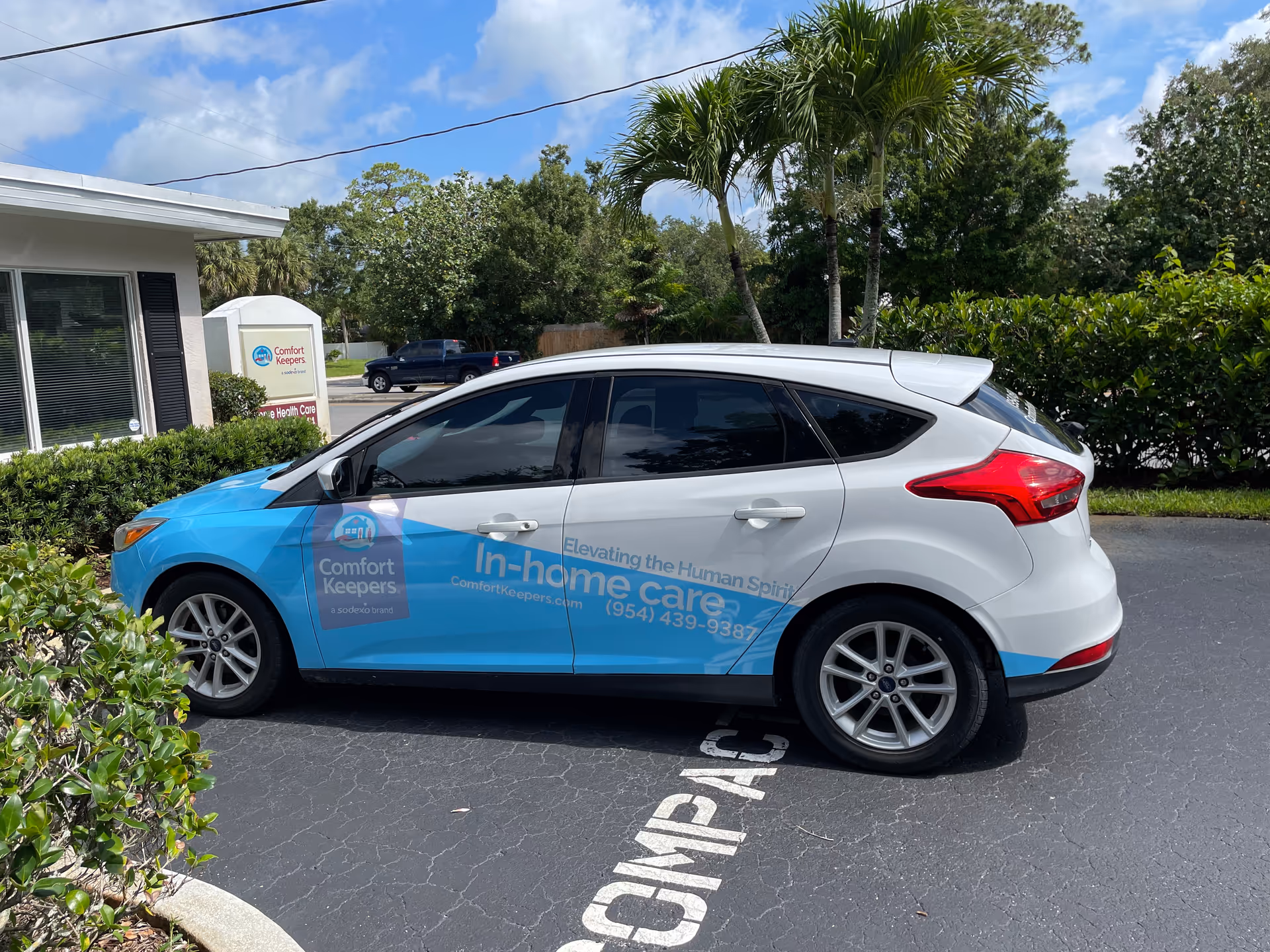Blue and white Comfort Keepers car parked in front of a senior care facility.