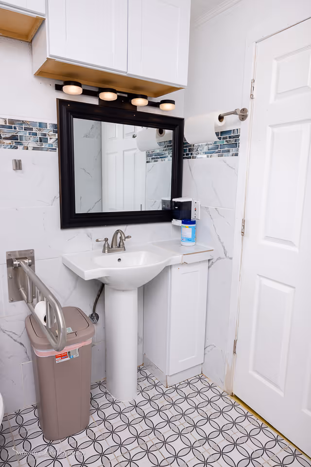 Small bathroom with pedestal sink, mirror, cabinet, trash can, and patterned tile floor.