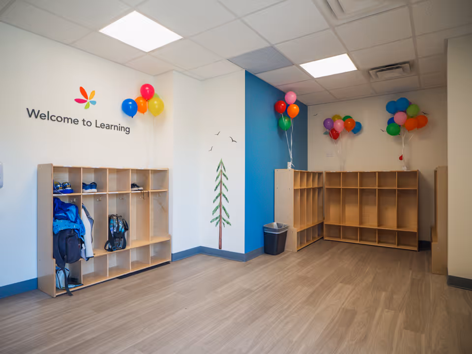 Bright interior learning room with cubbies, backpacks, and colorful balloons against a Welcome to Learning wall.