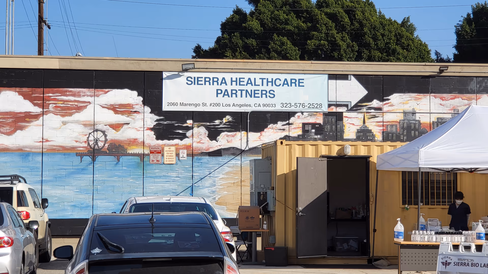 Outdoor scene of Sierra Healthcare Partners building with mural, banner, tents, and parked cars