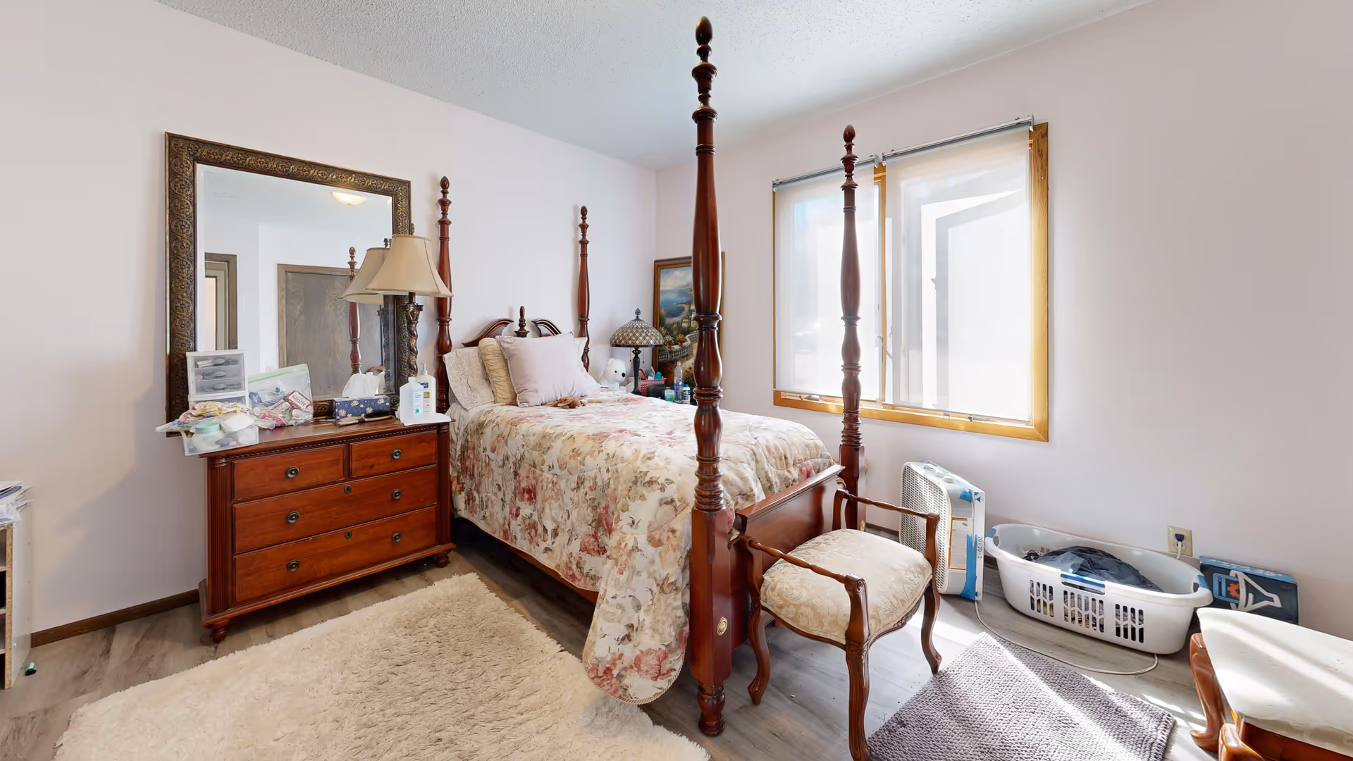 A bedroom with a four-poster bed, dresser, mirror, and floral bedding near a window.