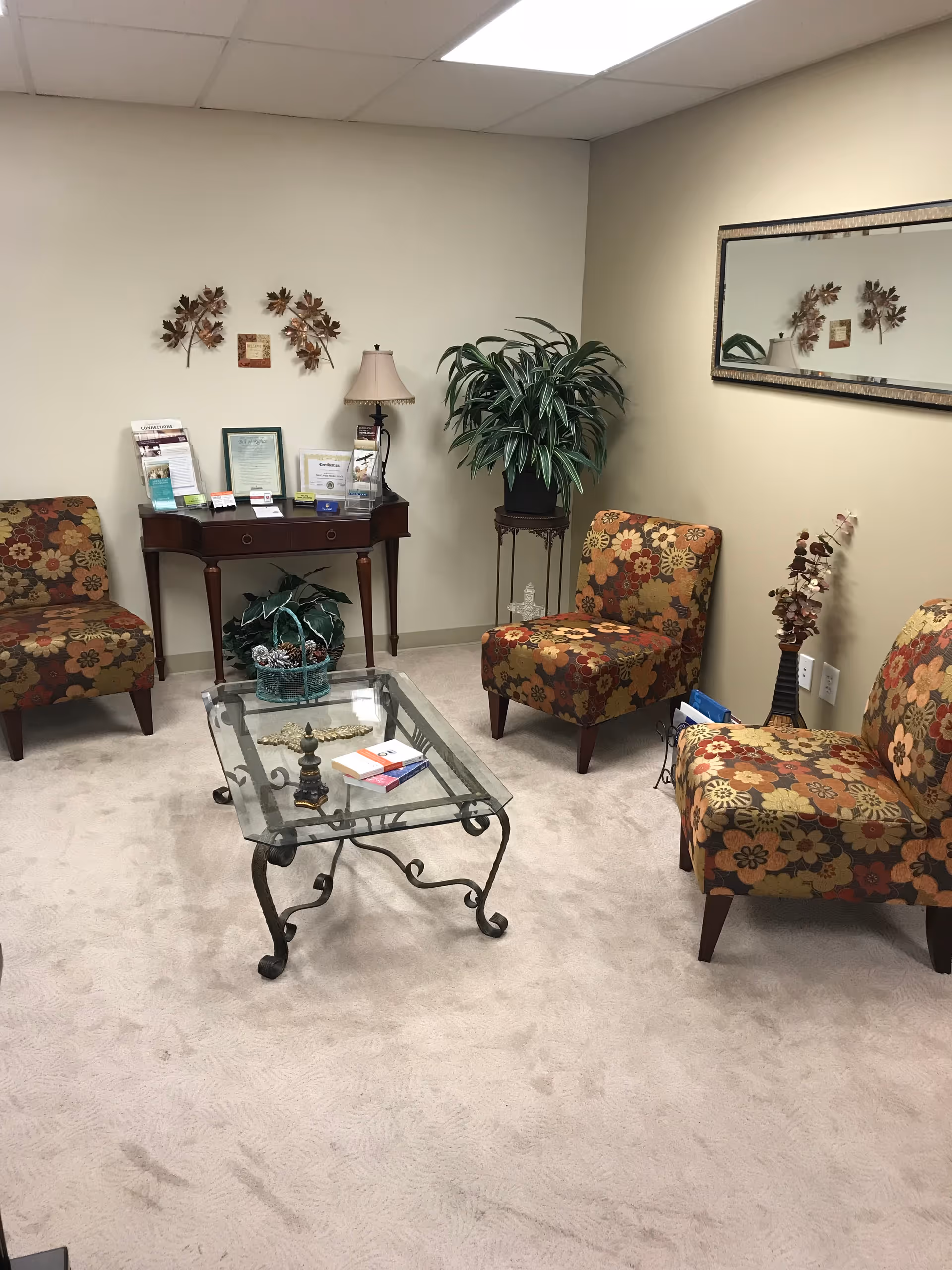 A cozy senior living lounge with floral chairs, glass coffee table, and plants.
