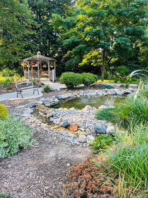 A landscaped garden with a wooden gazebo, pond, rocks, and benches.