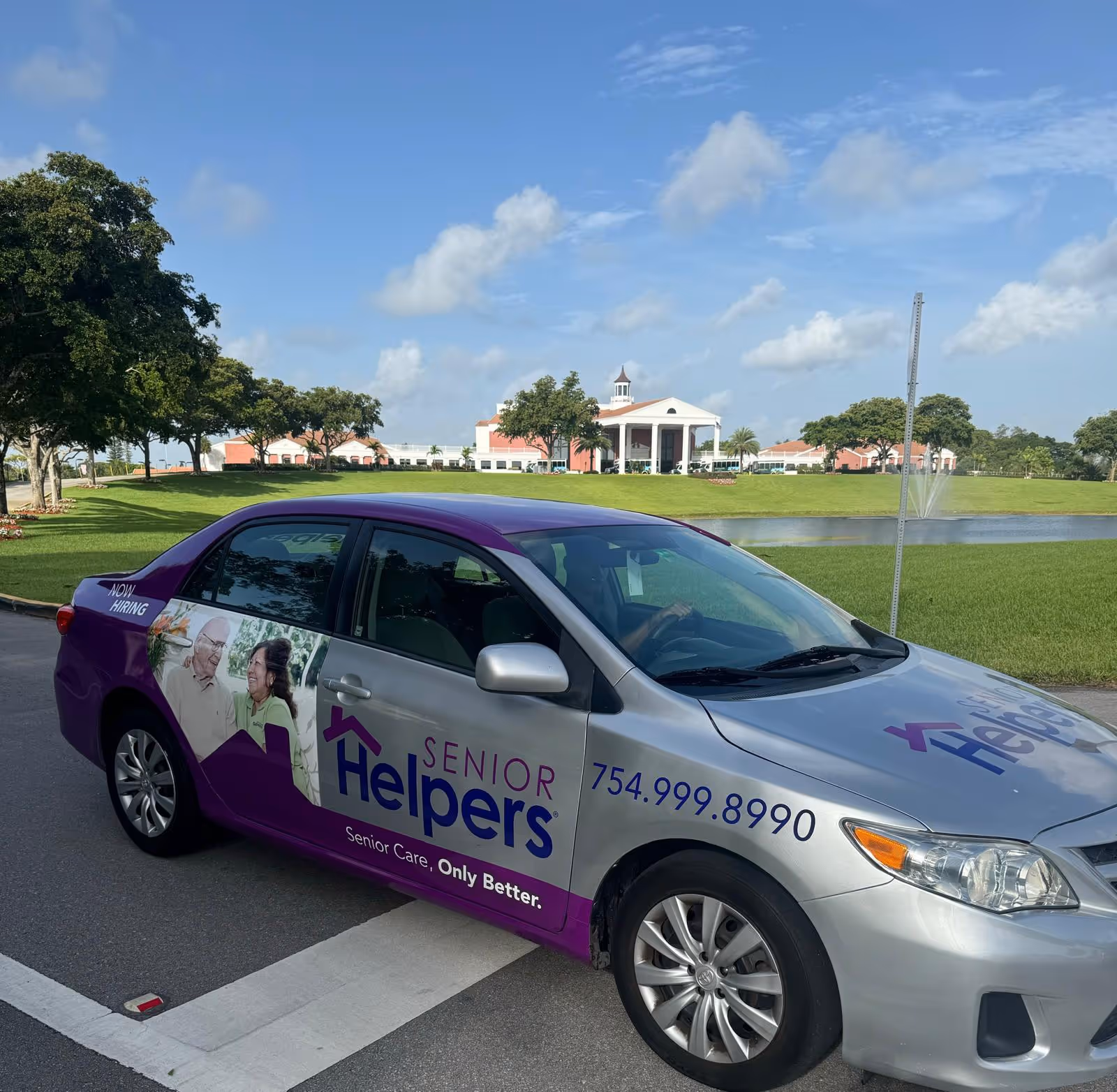 Senior Helpers branded car parked outdoors with campus building and fountain in distance.