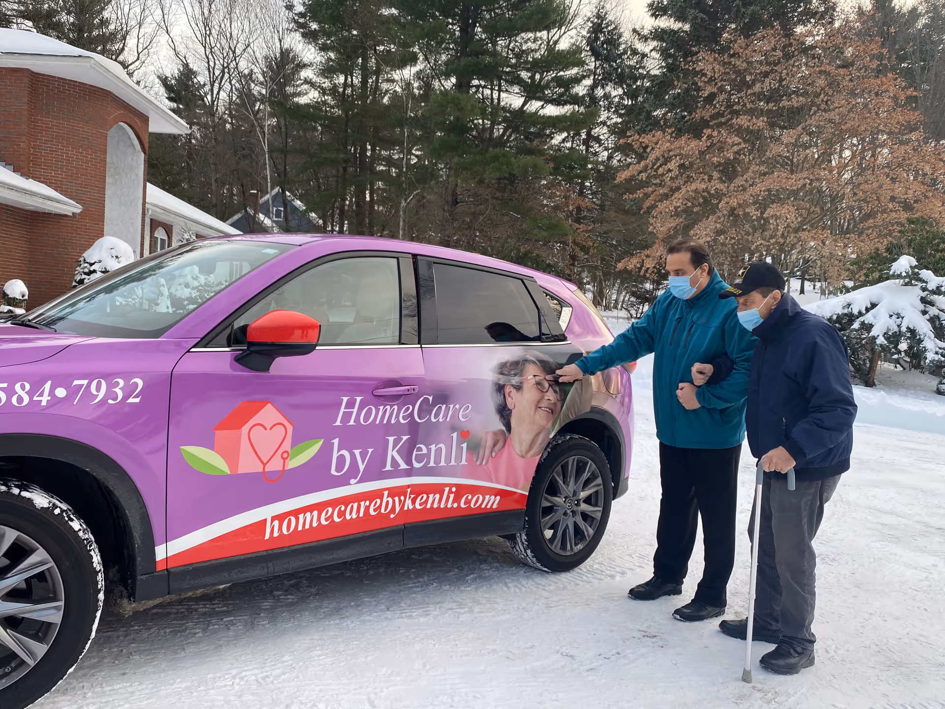 Two masked men stand beside a purple van labeled HomeCare by Kenli outdoors in snow.