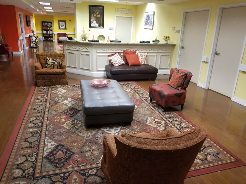 Cozy lounge with patterned rug, chairs, ottoman, and a front desk in a senior living facility.