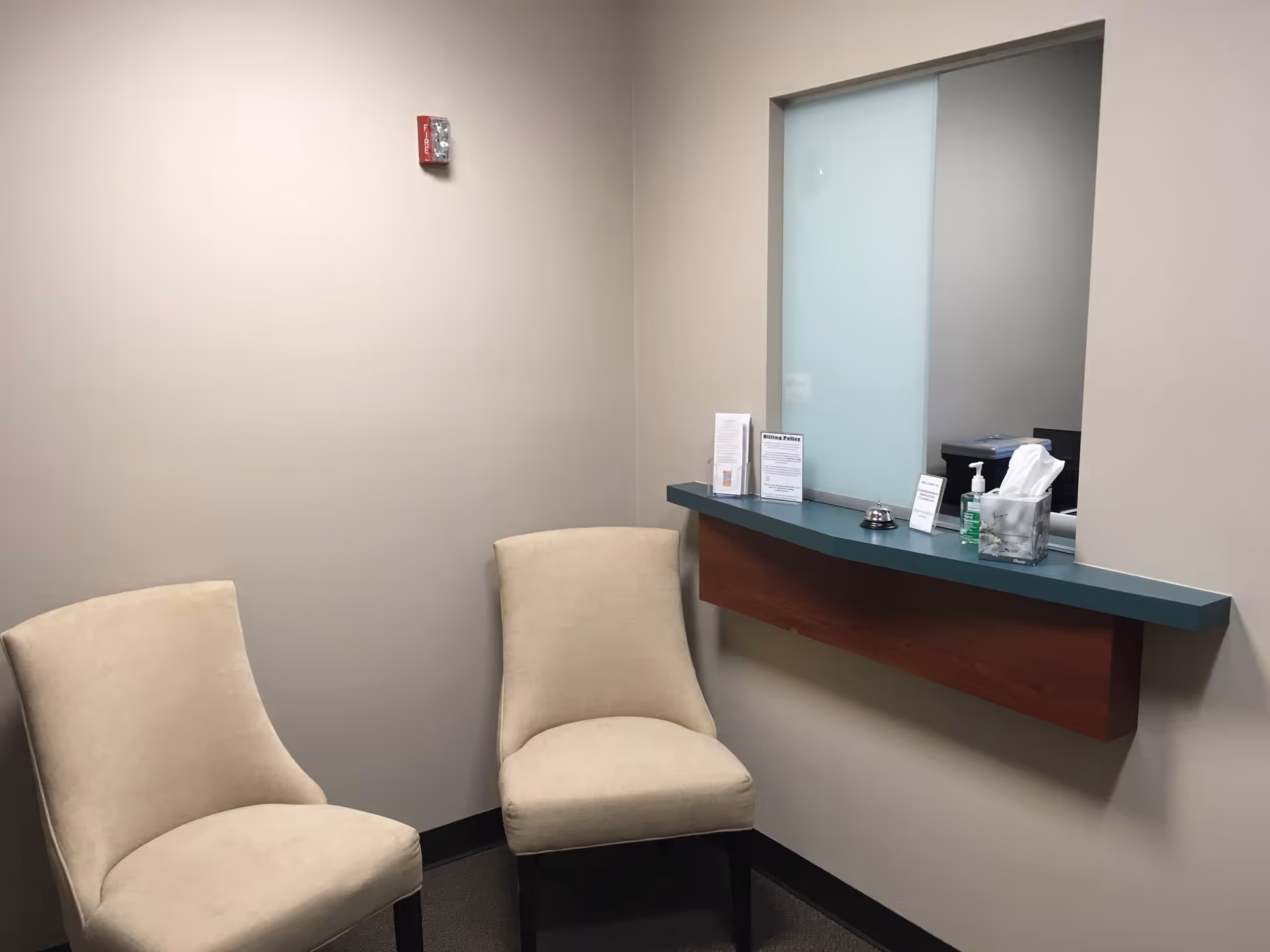 Two beige chairs sit beside a small reception counter with tissues and sanitizer.