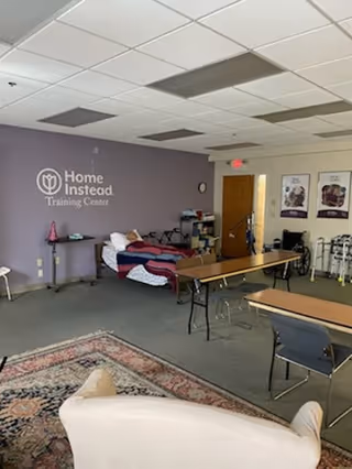 Interior training room with purple wall, Home Instead Training Center logo, desks, chairs, and a bed.