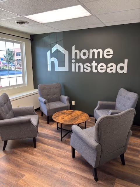 Lobby seating area with four gray armchairs around a round wooden table under Home Instead logo