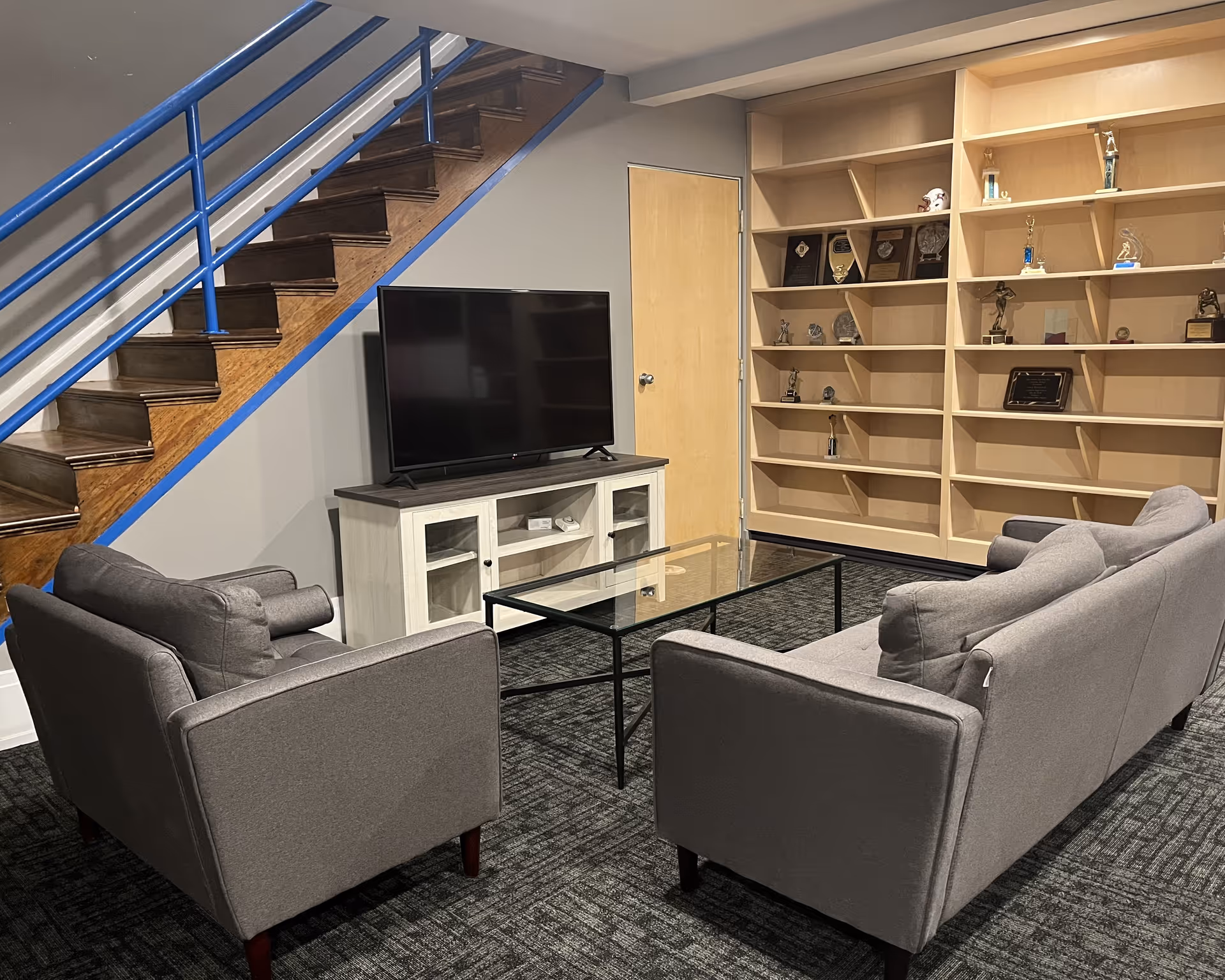 Common living room with gray sofas, glass coffee table, TV, and trophy shelves under a staircase.
